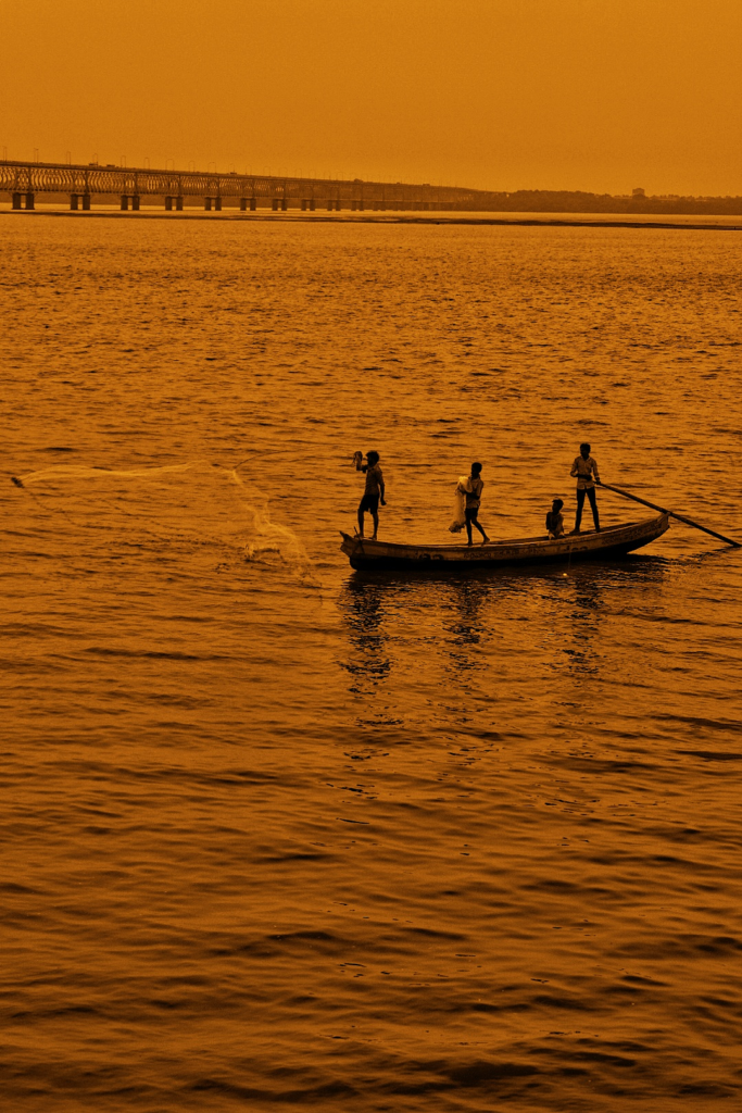 A group of people in a boat on the water, casting a net. A bridge is visible in the background.